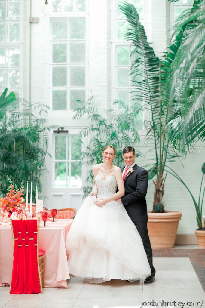 Bride in princess gown and groom standing by coral peach table with red chiffon fabric chairs and coral centerpiece